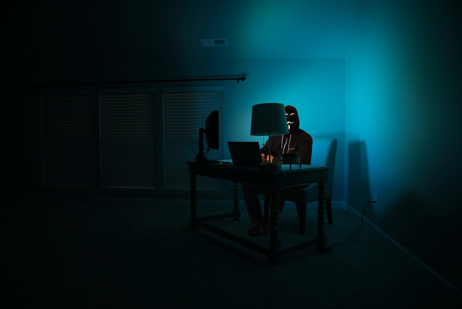 A hooded figure at a computer in a dark room